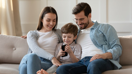 Smiling young parents with small preschooler son sit relax on couch in living room watch funny video on cell, happy Caucasian family with little boy child rest on sofa ta home using smartphone gadget