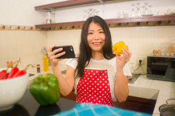 young happy and beautiful Asian Korean home cook woman in red apron reading healthy recipe in mobile phone cooking cheerful preparing delicious meal in kitchen