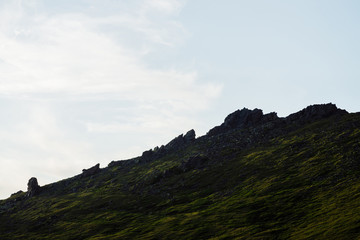 Silhouette of rocks in sunshine above cloudy sky. Vivid green mountain with crags in sunlight. Scenic minimalist alpine landscape in sunny day. Beautiful highland scenery with big stones on hillside.