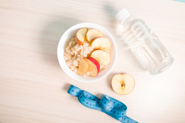 Fototapeta na wymiar half apple, bottle of water, a bowl of boiled oatmeal with sliced apple and a blue measuring tape on a wooden table, proper nutrition concept, healthy breakfast
