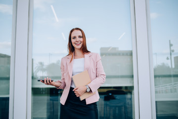 Prosperous redhead female manager holding copybook and mobile phone satisfied with successful career, portrait of smiling 20s business woman in elegant clothes looking at camera using gadget