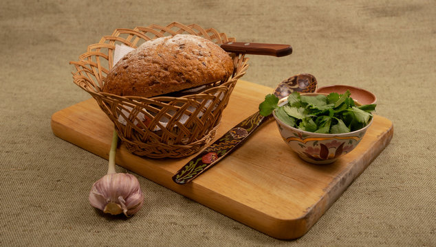 a whole round loaf of bread with a knife in a basket, a wooden painted spoon, garlic, herbs in a plate. wooden board