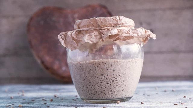fresh sourdough on whole grain flour in glass jar and rye artisan bread in shape heart, yeast-free leaven starter for healthy rustic vegetarian handmade bread, spices scattered on table