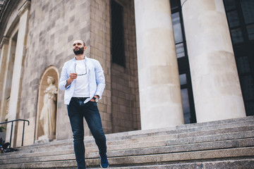 Confident hipster man taking off glasses while standing and relaxing on staircase of old building