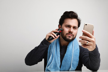 Bearded man with blue towel trimming his beard with a trimmer and using his smartphone watching video tutorials.