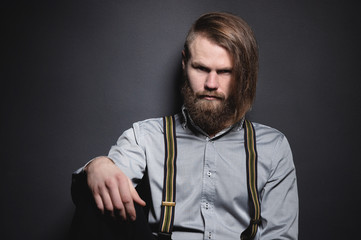 Contrasting low key portrait of a bearded long-haired stylish brutal man in a gray shirt with suspenders sternly looks at the camera. The concept of style and brutal beauty in the studio