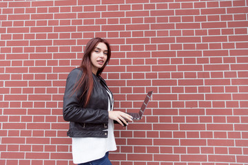Fototapeta na wymiar Red-haired girl in a black leather jacket works at the laptop against a red brick wall