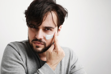 Fototapeta premium Close-up shot of attractive man with healthy skin, applies cream for anti wrinkle or anti aging, over white background.