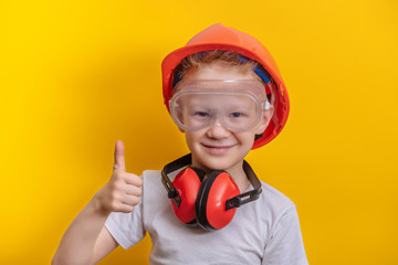 Cute boy in builder glasses, helmet and headphone to ear protection shown big finger up over yellow...