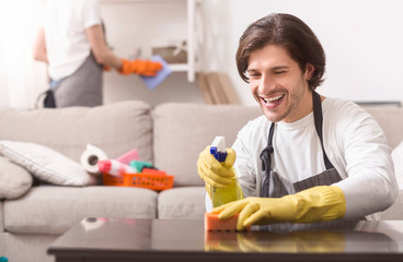 Domestic Cleaning. Smiling Man Wiping Dust From Table At Home