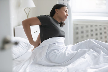 African mixed-race woman waking up sitting on bed feel low back pain after sleep caused by...