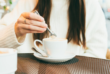 A young girl's hand puts sugar in coffee. On a round metal table there is a white cup of coffee and a young girl's hand puts sugar in it.