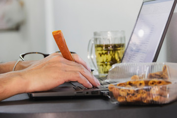 hand hold snack and  typing on keyboard of black laptop on desk at home office