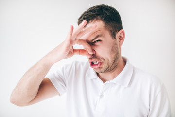 Young man isolated over white background. Guy covers his nose with fingers due to a bad smell. Look at camera with disgust.