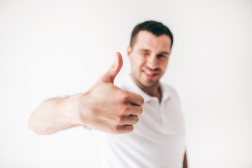 Young man isolated over white background. Guy hold one eye closed and show big thumb up. Like symbol. Cheerful attractive positive male person on blurred background.