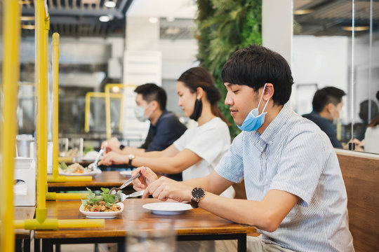 Asian man and woman sitting separated in restaurant eating food with table shield plastic partition to protect infection from coronavirus covid-19, new normal restaurant and social distancing concept