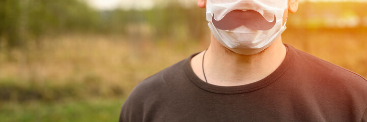 father's day during the covid-19 quarantine concept. portrait a young european man in a protective mask and paper moustache against the background of nature. flare. banner