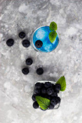 A glass with a blue drink and blueberries with mint leaves with water drops on the surface. Next to it is a plate of blueberries lying on a white table with green leaves of Melissa