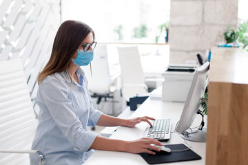 Obraz na płótnie Canvas Young woman wearing medical mask in office. Protection employees on workplace. Girl working at reception indoors. Social distancing in public place, disease prevention during quarantine, staff safety.