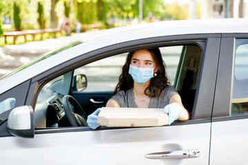 Safe food delivery from pizzeria to car during quarantine covid-19. Attractive young girl in blue medical mask and gloves gets pizza in cardboard box. Delivering food by car according social distance.