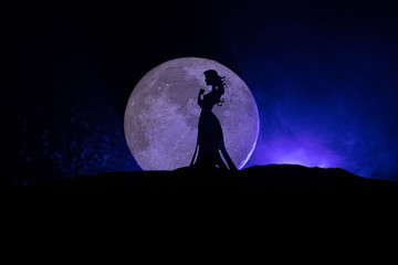 Girl standing alone behind big full moon. Dark toned foggy background. Loneliness bad mood concept