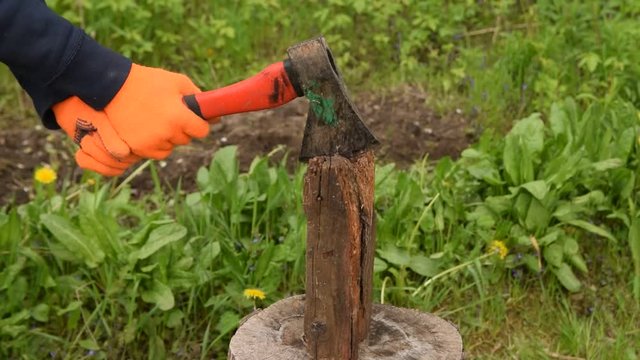 Hands in orange gloves hold an ax with a red handle and chop wood on a stump