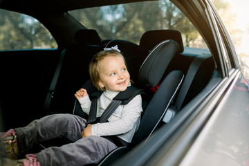 Beautiful smiling cute baby girl fastened with security belt in safety car seat, looking at the window. Child transportation safety concept