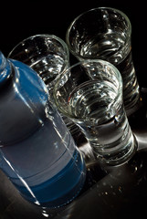 Glasses with alcohol near the bottle. Transparent drink on a black background. Blue reflexes on the bottle.
