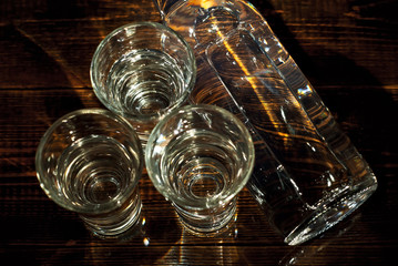 Glasses with alcohol near the bottle. Transparent drink on a brown wooden background. Table made of dark shabby boards. Blue reflexes and bright reflections of light on the bottle.