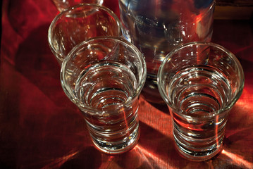 Glasses with alcohol near the bottle. Transparent drink on a red wooden background. Blue reflexes on the bottle.