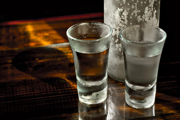 Frozen glasses with alcohol near the bottle. Drinks on a dark brown table. Transparent utensils on a black background. Old shabby boards under glass.
