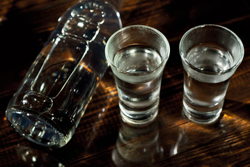 Frozen glasses with alcohol near the bottle. Drinks on a dark brown table. Transparent utensils on a black background. Old shabby boards under glass.