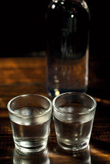 Frozen glasses with alcohol near the bottle. Drinks on a dark brown table. Transparent utensils on a black background. Old shabby boards under glass.