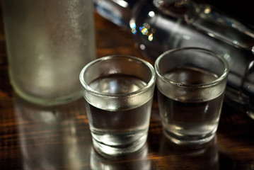 Frozen glasses with alcohol near the bottle. Drinks on a dark brown table. Transparent utensils on a black background. Old shabby boards under glass.