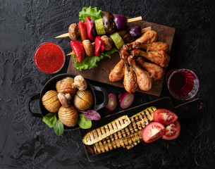 Fried chicken legs, baked potatoes and vegetable skewers on a black table. The view from the top.