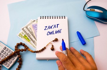 Note book with texts zakat online (alms-giving), calculator, beads and mouse on blue. Conceptual of  payment during Covid-19 lockdown. Muslim obligation to aid the poors. Selective focus.