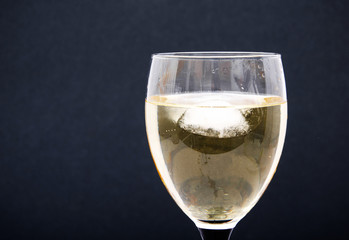 A glass of white wine with pieces, ice cubes. Wine with ice on a black background. Glass with a black  leg