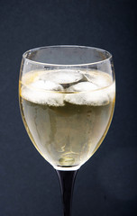 A glass of white wine with pieces, ice cubes. Wine with ice on a black background. Glass with a black  leg