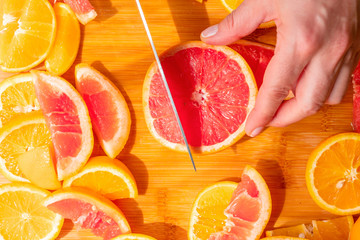 Top view of hand holding a fresh grapefruit slice and cutting with a knife new pieces. Half and circle shape sliced oranges and citrus fruits lying flat on a wooden board