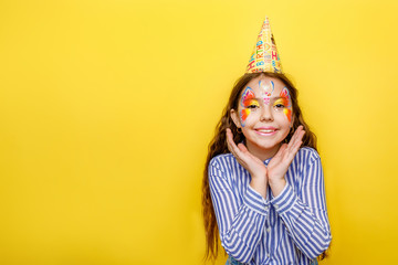 Little girl in birthday party hat hands spread up screaming isolated on a yellow background. Place for text, copy space