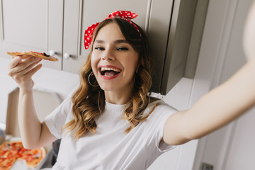 Refined lady with waving hair making selfie during dinner. Indoor shot of blissful caucasian girl eating pizza.