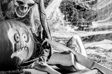 Black and white photo with strong grain effect. Laughing skeleton who is looking at scared pumpkin which is looking on other sliced pumpkin and hand holding the knife. Halloween,  artistic concept.