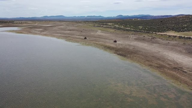 A pair of ATV racing, running, on dirt along the shoreline of a reservoir raising puffs of dust as they go with mountains in the background and overcast sky