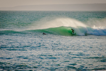 Surfer in the barrel of a turquoise wave, spray behind and another surfer watching while paddling out, evening light