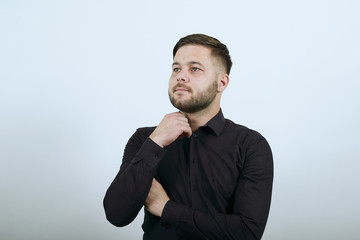 Young Bearded Dark Haired Man In Black Stylish Shirt On White Background, Dreamy Male Holds Hand To His Chin And Thinks. The Concept Of Smart People, Intellectuals