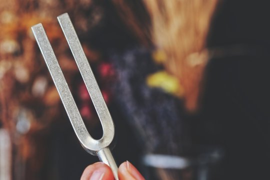 Close up of female holding a tuning fork in her hand in between fingers. Super blurred colorful background with purple, blue, pink, yellow dried flowers and herbs. Silver metal 741 hz sound fork 