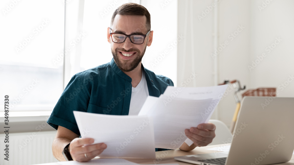 Wall mural happy handsome businessman holding and reading documents with good news near laptop. young smiling b