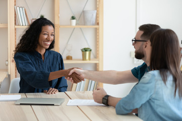 Real estate agent shaking hands with happy young couple in office at meeting. Smiling millennial family clients take congratulation on buying house rent and mortgage by financial advisor.