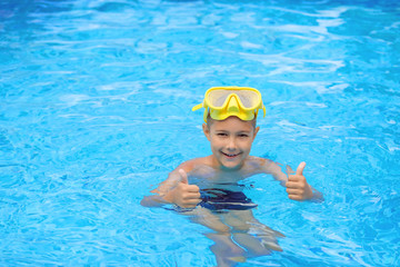 A child in a mask for swimming. A young boy in the pool shows thumbs up. Kid are engaged in diving.