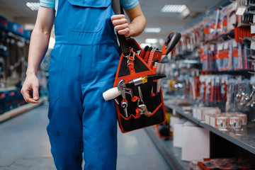 Male worker in uniform holds toolbox in tool store - obrazy, fototapety, plakaty
